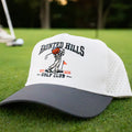 Vintage Style Golf Hat Halloween Golf Hat gift idea for golf dads. A white and dark gray trucker hat with 'HAUNTED HILLS GOLF CLUB' printed on the front, resting on a green golf course with a golfer blurred in the background.