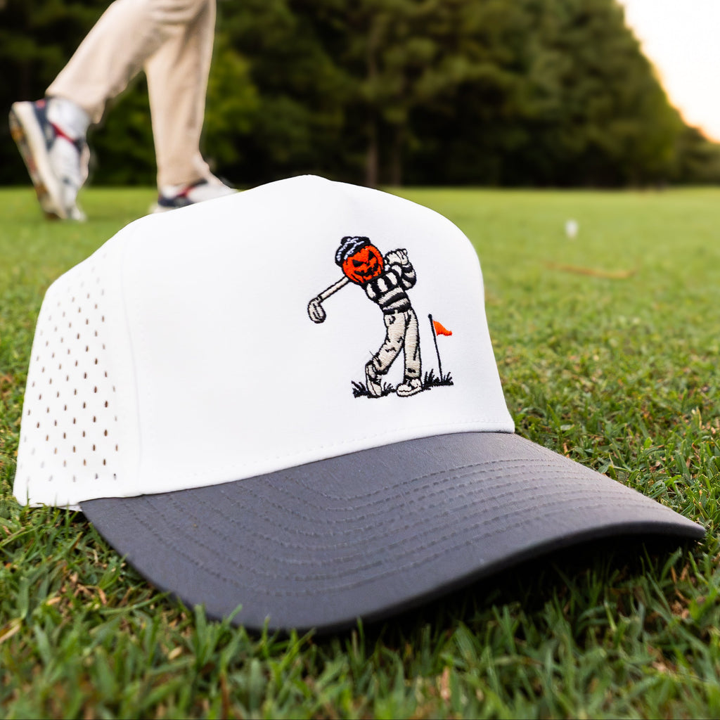 Vintage Style Golf Hat Gift Idea. Halloween Golf Hat gift. A white and navy blue trucker hat with a cartoon golf ball player graphic on the front, resting on a green grass golf course. A blurred golfer is visible in the background.