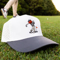 Vintage Style Golf Hat Gift Idea. Halloween Golf Hat gift. A white and navy blue trucker hat with a cartoon golf ball player graphic on the front, resting on a green grass golf course. A blurred golfer is visible in the background.